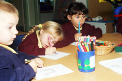 Three young children focus on writing in a school environment. Each has their own paper and there is a jar of coloured pencils on the table in front of them.