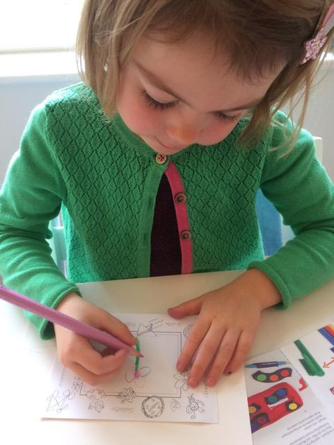 A girl draws and colours a picture of a flower with her right hand, steadying the paper with her left. 